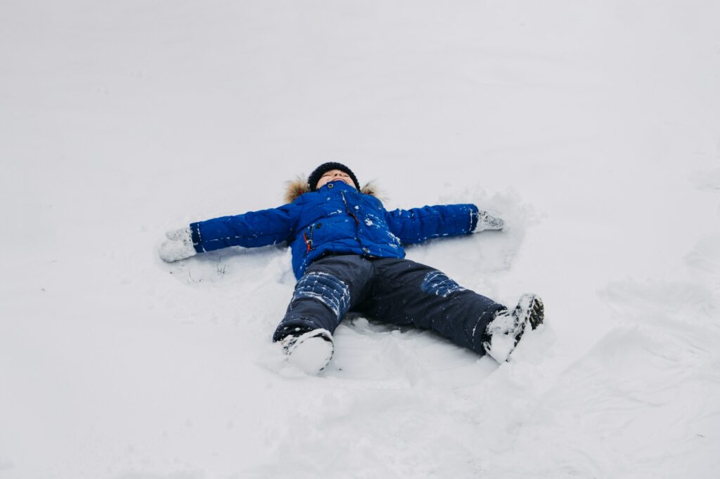 Best Outdoor Winter Activities for kids. Little kid boy in blue winter jacket making snow angel